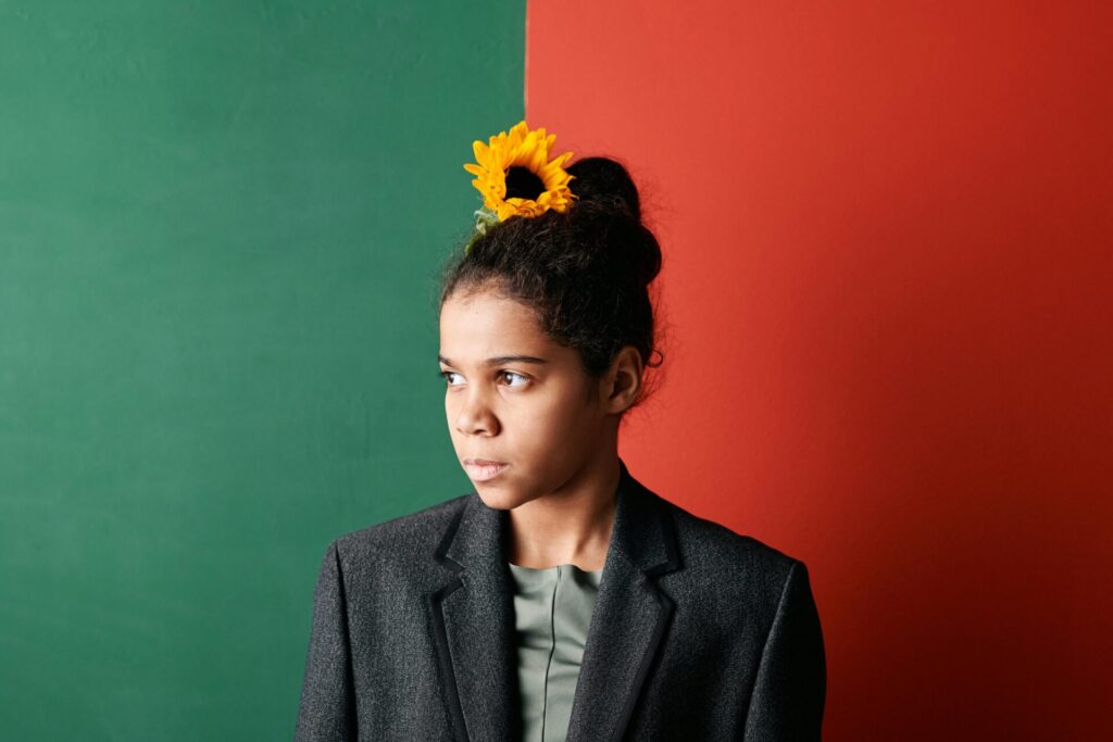 Portrait of a young black woman in a blazer with a sunflower in her bun against a red and green backdrop.